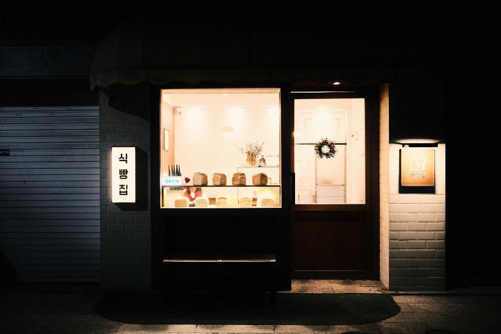 A small bakery at night. One lit window with bread in it, an open sign, shutters down either side.